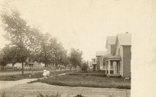 View across road towards a neighborhood along another road. Houses are on the right, and two children in the center are standing on the sidewalk in the center.