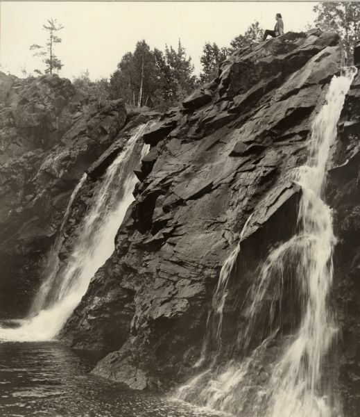 Little Manitou Falls on the Black River. Referred to as the "upper falls" in the park. A person is sitting on the rocks on top of the falls. This waterfall has a drop of some thirty feet. The park was named for its donor, Martin Pattison.