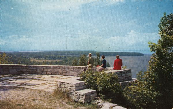View looking west from Eagle Bluff toward Nicolet Bay, the Strawberry Islands, and Chambers Island. Three people are sitting on the stone wall of the overlook for a view towards the bay.