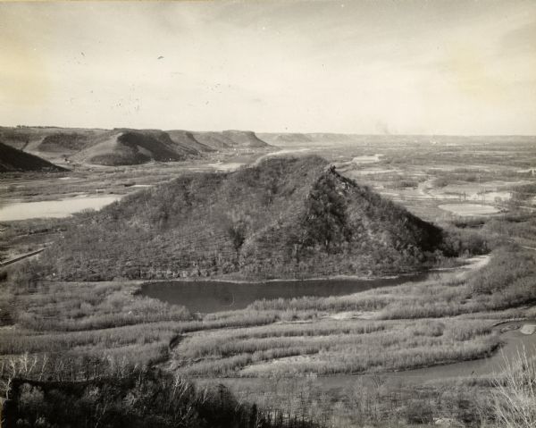 Aerial view looking east at Mt. Trempealeau from Brady's Bluff in Perrot State Park. The Mississippi River is visible along the bluffs at upper left and the Trempealeau River is visible in the foreground. The name "Trempealeau" was given to this land-form by seventeenth century French explorers. It is a (romanticized) transliteration of the Winnebago and Chippewa names for the "Mountain which bathes its feet in the water".