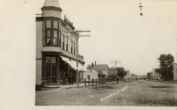 Road in town lined with commercial buildings on the left side. Caption reads: "Pittsville, Wis."