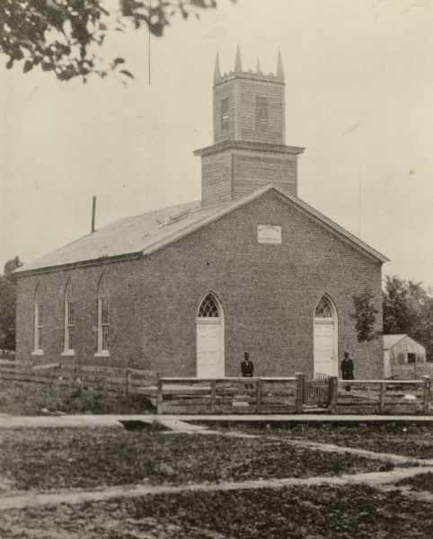 A view of the First Methodist Church. Two men are standing in front of the church behind a fence.
