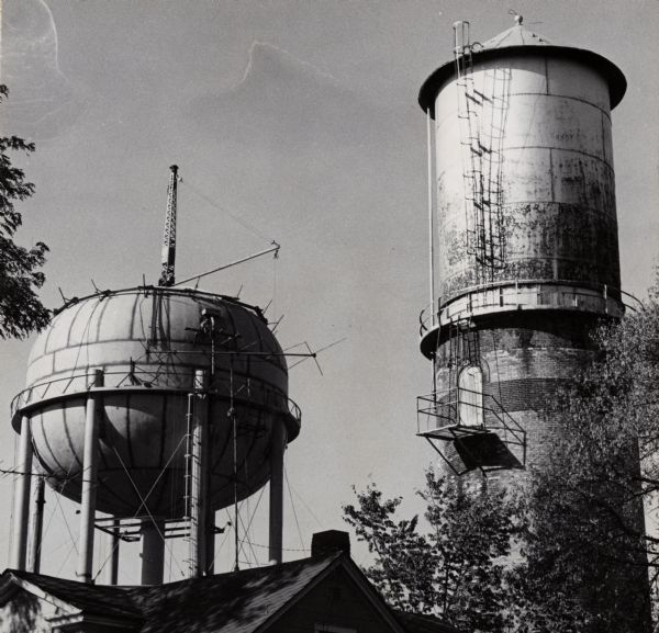 View looking up at two water towers. The roof of a building and treetops are in the foreground.