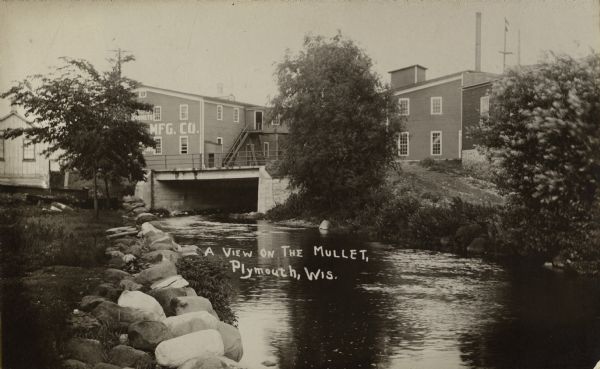 View of the Mullet River, with an bridge and an industrial building in the background. A sign reads, in part: "MFG. CO."