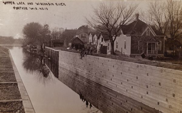 The Portage Canal at the Wisconsin River, showing the upper lock. Caption reads: "Upper Lock and Wisconsin River Portage Wis. No. 10". A group of people are sitting and standing at the top of the opposite wall. Houses are in the background.