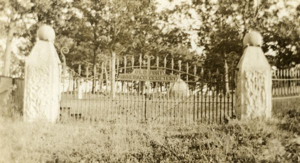 View through the closed gate, which has a sign that reads: Old Fort Winnebago Cemetery. Overgrown plants surround the gate, and trees surround a monument just inside the gate.