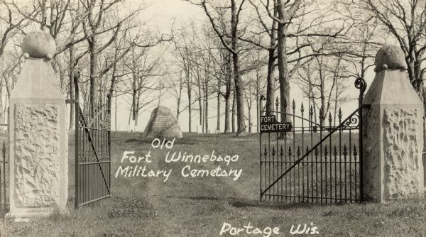 Caption reads: "Old Fort Winnebago Military Cemetery". View through the open gates of the cemetery. Trees surround a monument just inside the gates.