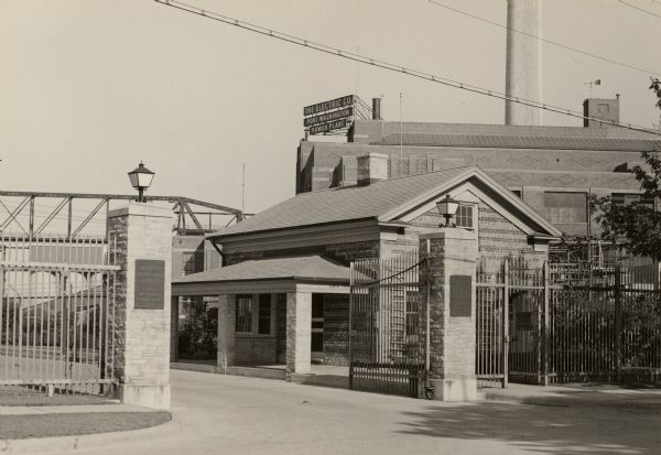 A view of the Pebble house, also called Edward Dodge House. Built in 1848 and constructed of cobblestone. Stone columns with a gate and fence are in front of the house. Plaques on the columns read: "Wisconsin Electric Power Company — Port Washington Power Plant". A large industrial building in the background has a sign that reads: "The Electric Co. Port Washington Power Plant".