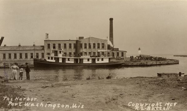 View towards the shoreline of the harbor. A man, woman and child are standing at the shoreline on the left. Across the water is a boat along the opposite shoreline near an industrial building with a smokestack. Caption reads: "The Harbor, Port Washington, Wis."