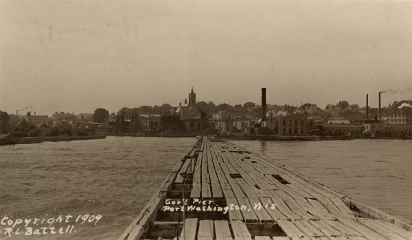 View down Government Pier. In the distance are industrial buildings along the shoreline. Caption reads: "Gov't Pier Port Washington, Wis."