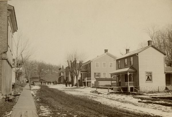 View down sidewalk on the left side of an unpaved street. A brick building is in the foreground on the left. Dwellings are along the right, and snow is on the ground. In the distance is a tree-covered hill.