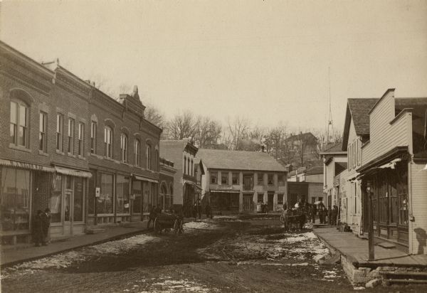 View down unpaved street of lined with commercial buildings. Pedestrians are on the sidewalks, and some snow is along the curbs. In the background is a hill with a building on top surrounded by bare trees.