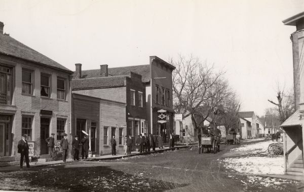 View down unpaved street with snow along the curbs. The Post office is the first building on the left. Other buildings are occupied by a doctor's office, saloon, livery and feed stable, etc. Men are standing along the sidewalk on the left. Persons identified by name, but no identification of placement, are: Wes Hymer, O. Seaton, Louie D Reinieke, Dr. McGovern, H.E. Coons, Andy Kaltenbach, J. Henry Coons, George Chalders, John Busher, and Henry Doser (white shirt).