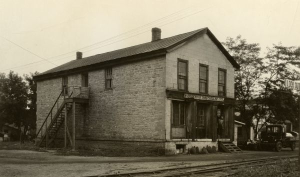 Former building of the Astor Company, also known as the Brisbois Fur Trading Post. At the time this photograph was taken, the structure was occupied by Ben Schaub's Riverside Repair Shop. A man is standing at the entrance.