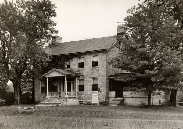 Exterior view of the Brisbois House. A sign is on the right side of the entrance, and reads, in part: "Brisbois House Home of Michael Brisbois Early Fur Trader".