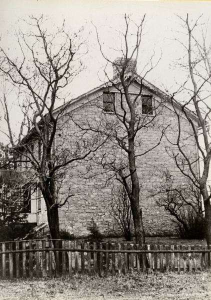 View of the Brisbois House, east elevation. A fence and trees are in the foreground.