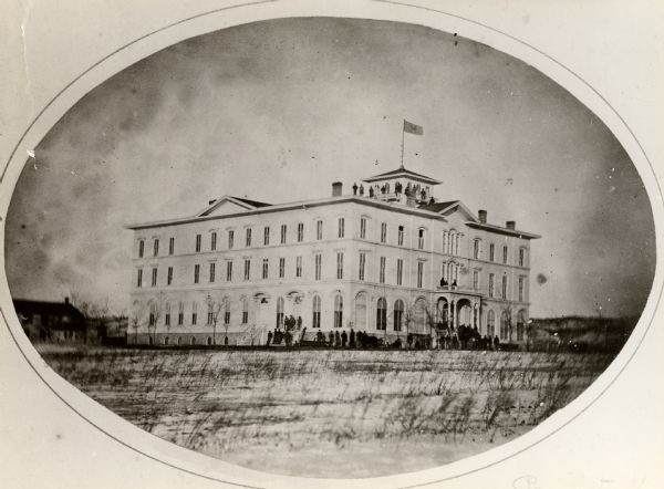 Oval-framed view of Lawler Hall at Campion College. This image was made during the Civil War when the building was used as a U.S. Military hospital. Prior to the war, it was Brisbois Hotel, built in 1857. A group of people are standing around the front and sides of the building. Another group of people are posing on the roof.