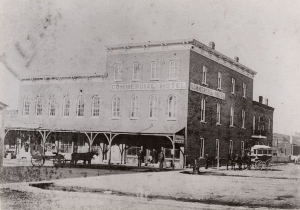 Exterior view of the Commercial Hotel. Horse-drawn vehicles are in front of the building. People are standing on the sidewalk in front.