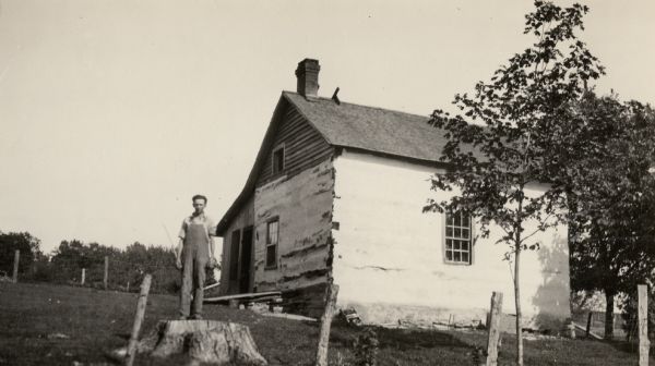 A view of the DeLury log cabin, where four generations (at time of photograph) of the same family have lived. Probably built around 1840. A man is standing on the stump of a tree in the foreground.