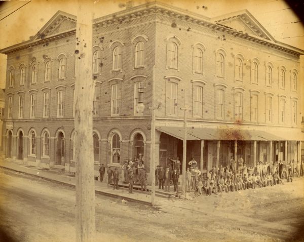 Elevated view across street towards the Dousman Office building, later used as a pearl button factory. A large group of people are sitting and standing at the corner of the building, some on the sidewalk on the left, and more under the porch along the street on the right.