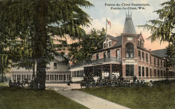 Exterior view of the Prairie du Chien Sanitarium. Established in 1903, it became a general hospital in the 1930s. A group of people are posing on the lawn and the porch of the building. Caption reads: "Prairie-du-Chien Sanitarium, Prairie-du-Chien, Wis."
