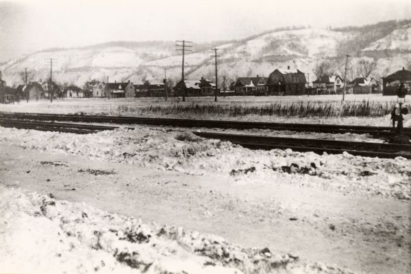 View of the Burlington Railroad tracks south of the city, near Campion College. Dwellings are across a field, and snow-covered bluffs are in the background.