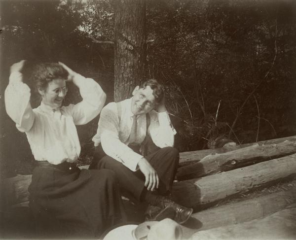 Young woman and man, a Mr. Lyman Sinclair, sitting on a log against a tree. The man is smiling and resting one elbow on his knee, while running his hand through his hair. The woman is sitting on the left, wearing a long skirt and shirtwaist, and is looking down while fixing her hair. There is a hat in the foreground.