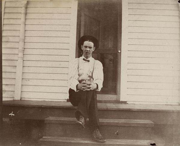 Oscar (Boney) Halverson smoking a cigar on the front porch of a house. He is wearing a shirt with collar, bow tie, suspenders, and high, laced boots, and is sitting on the top step with hands gripping his crossed legs, and his hat pushed back on his head.