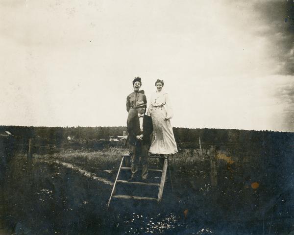 A man and two women standing on a stile of broad wood in the middle of a field. The man standing on the second step, wearing a coat, bow tie, and fedora. The two women are standing behind him on the top step, wind blowing their skirts. The woman on the left has her mouth wide open. Halycon Brick Yard is in the background.