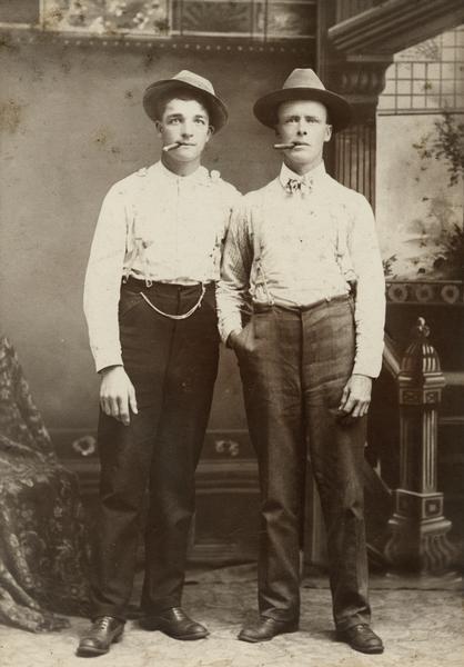 Studio portrait of Jake Hubert (wearing the watch chain), and another man, both smoking cigars and wearing hats and standing in front of a painted backdrop.