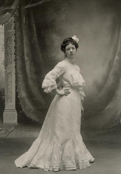 Young woman facing to the right with her head turned toward viewer, hand on hip, wearing wire-rimmed eyeglasses, a flower in her hair, in long white dress with ruffles at bodice and hem, a slight train, puffy sleeves gathered at wrist, standing before a painted studio backdrop of drapery and a pillar. Possibly in a wedding gown?