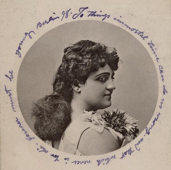 Head and shoulders portrait from right side, with head turned partially forward but not looking directly at viewer. She has long hair gathered at neck and wears a large corsage and theatrical make-up. Image is cut to oval with handwriting in blue ink around perimeter: "To things immortal time can do no wrong, and that which never is too <i>[sic]</i> die - forever must be young. Berlin '78."