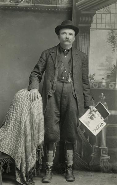 Studio portrait of William Tennant in front of a painted backdrop. He was a newspaper and magazine agent in Black River Falls, Wisconsin, who lost both his legs below the knee. He is facing the viewer, with his right hand on a draped chair, and in his right hand he is holding a magazine. His pant legs have been shortened to reveal his artificial legs, and is wearing a suit and derby hat.