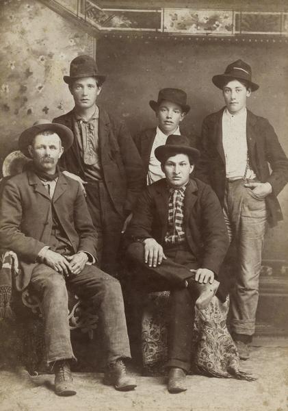 Group portrait of five men in front of a painted backdrop. Two men are sitting; all the men are wearing hats.