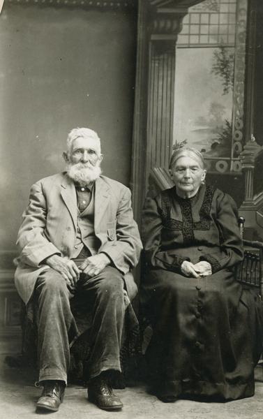 Studio portrait of an elderly couple, both seated in chairs in front of a painted backdrop.