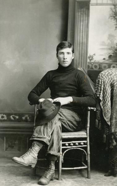 Studio portrait of young man sitting holding a hat in front of a painted backdrop.