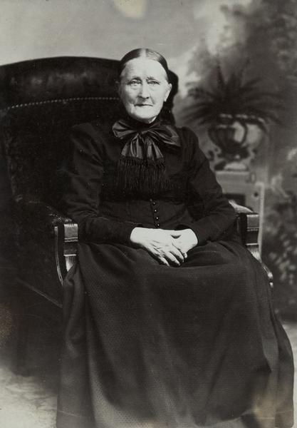 Studio portrait of elderly woman in black dress with large bow at neck, seated in chair with hands crossed in her lap in front of a painted backdrop. She wears eyeglasses and looks directly at viewer.