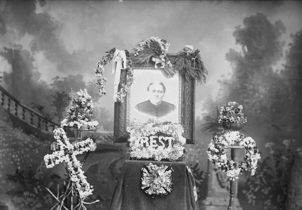 Framed woman's portrait surrounded by funeral wreaths and flower arrangements, with painted studio backdrop. A is cross on the left, with an arrangement spelling "rest" in front.