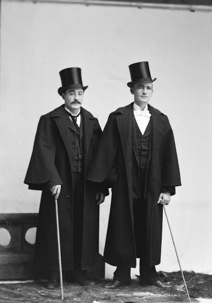 Studio portrait of two standing men wearing greatcoats and top hats, and holding canes. The man on the left is probably John Carish, who worked in the butcher shop, and on the right, Edward Oderbolz, the son of the brewer who later drowned in a beer vat.