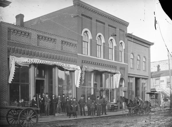 Seventeen men and boys are posing in front of the storefronts on a sidewalk on the north side of Main Street, between First and Second Streets, including O.C. Hearn Grocery, Chicago Cheap Store, and Moe's Hardware (with a sign reading "Garland Stove Ranges"). The wagon pulled by two horses on the right is probably a photographer's wagon.