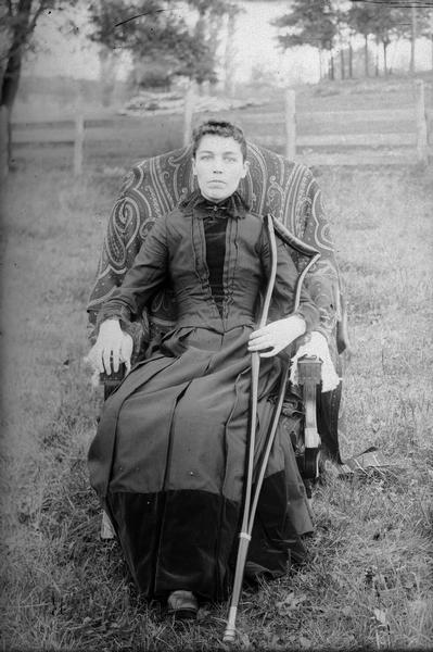 Woman posing sitting in a chair and holding a cane in the yard of a house, probably Mrs. Hassel Phillips. 
