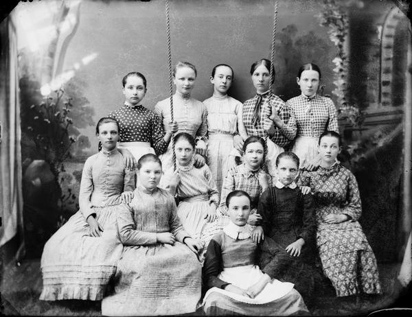 Studio portrait of twelve young women posing, seven sitting and five standing, around and on a swing in front of a painted backdrop. Seated at left in the first row is probably Jennie Gerioux White, and sitting in the second row, second and third from the left, is possibly Julie Jones Comnock and Margaret Murray Sample.