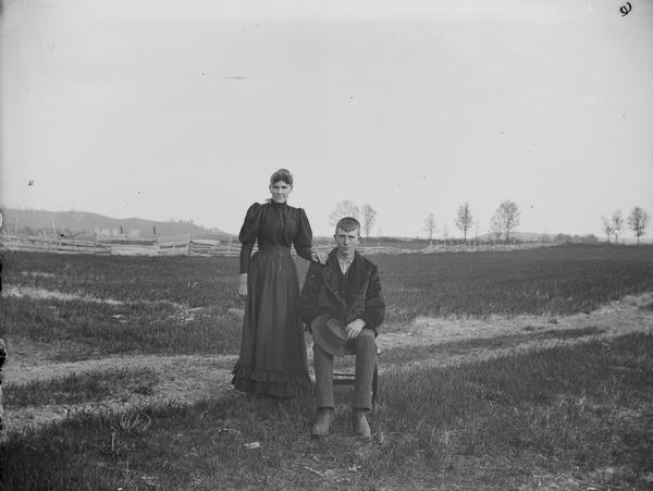 Man sitting on chair, and woman standing with hand on his shoulder, in a field with a wooden fence in the background. He holds a hat in his hands. She is dressed in dark mutton sleeves with lighter skirt, piped at hem.