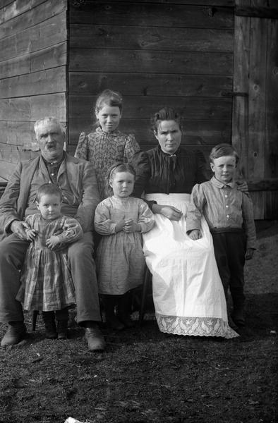 Old, bearded man and younger woman sitting in front of the corner of a wood outbuilding surrounded by four young children. A young boy is standing at his mother's side on the right, an older girl is standing behind the man and woman, a younger girl is in the middle. The youngest girl is standing between the older man's legs. The woman is wearing a dark blouse and long, white apron.