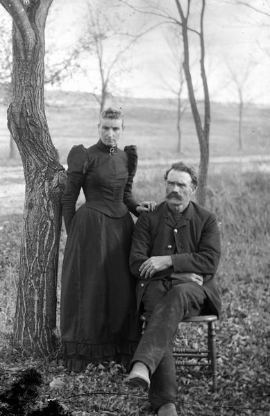 Man posing sitting in a chair, and a woman posing standing next him, with a tree on the left.