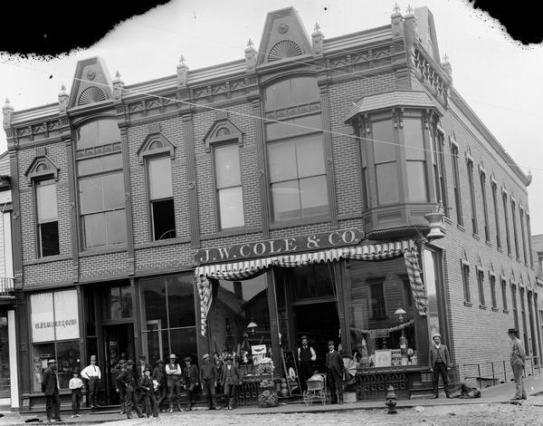 Sixteen men and boys posing in front of the building holding the J.W. Cole & Co. Drugstore and W. Elmore & Sons Store on the northeastern corner of Water and Main Streets.  Man near store entrance stands beside baby buggy.