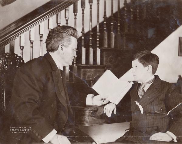 Posed portrait of Senator Robert M. La Follette Sr., and Jr., seated at a table with a stairway behind them.  It is likely this photograph was taken in the La Follette's Washington, D.C. home.
