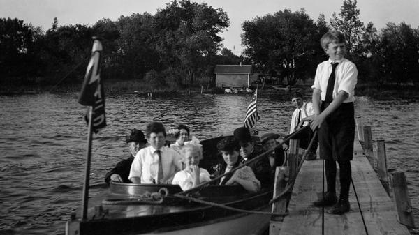 The La Follette Family in a boat, probably on Lake Mendota, with Gilbert Roe, La Follette's law partner, and his wife.  Senator Robert M. La Follette, Sr., is seated to the far left wearing a dark hat.  From the left the others are Bob, Jr.; Netha Roe; Mary La Follette; Belle Case La Follette; and Gilbert Roe.  Philip La Follette is standing on the dock holding the rope.  The other person on the dock is identified only in the La Follette Family Album as "Joe."