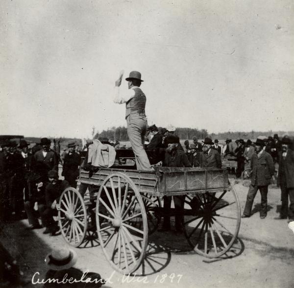 Robert M. La Follette, Sr., with his fist in the air, speaking from the back of a wagon. He is wearing a hat and a suit without the coat. It was in part due to his vigorous speaking style that La Follette won the nickname "Fighting Bob." This image is one of a series of views of his appearance at a fair in Cumberland, Wisconsin, in 1897. After three unsuccessful campaigns during which he brought his reform message to Wisconsin at events such as this, La Follette was elected governor of Wisconsin in 1900.