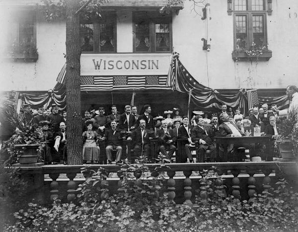 The official Wisconsin delegation photographed at the Wisconsin Exhibit at the St. Louis World's Fair in 1904.  Seated at the table, (at the right) are Governor Robert M. La Follette, Sr., and Lieutenant Governor James O. Davidson.  Former governor William Dempster Hoard can be partially seen under the flag to their right.